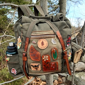 May include: Olive green canvas backpack with brown leather straps and patches. Patches include a bee, leaves, and flowers. A water bottle is in the side pocket. The backpack is hanging on a tree branch.