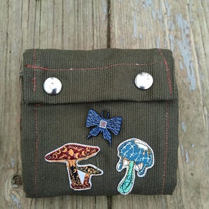 May include: Olive green canvas wallet with two silver snap closures. The wallet is decorated with three embroidered patches: two mushrooms and a blue butterfly. Red stitching accents the edges of the wallet.