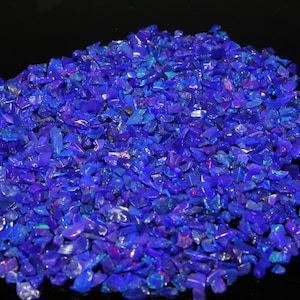 Op de afbeelding: Een stapel verpletterde, levendige blauwe opaal edelstenen. De opalen hebben een mix van vormen en maten, met kleurflitsen. De edelstenen zijn verspreid op een zwart oppervlak, wat een opvallend contrast creëert.