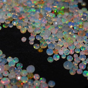 May include: A collection of small, round, faceted opal gemstones featuring a translucent white base with vibrant flashes of green, red, orange, and blue light. These loose, polished stones vary in size and display a high-gloss finish.