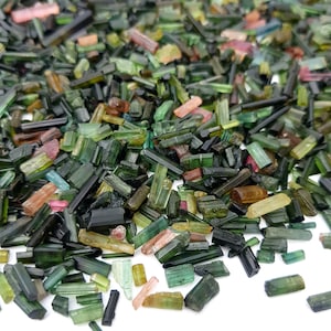 May include: A pile of raw tourmaline crystals in various shades of green, pink, and black. The crystals are small, rectangular, and have a natural, unpolished appearance. The crystals are scattered across a white surface.