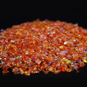May include: A close-up of a pile of orange opal chips. The chips have a sparkling, iridescent quality with flashes of green, purple, and yellow. The gemstones are small and irregular in shape, with a dark background.