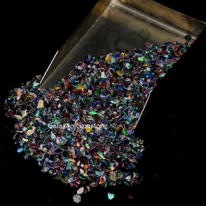 Peut inclure: Un ensemble d'opales concassées iridescentes, affichant un spectre de couleurs comprenant le bleu, le vert, le rouge et le jaune. Les opales se déversent d'un plateau métallique sur une surface noire. Le texte "Galaxy of Gemstone" est visible.
