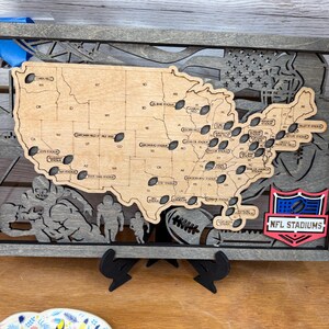 May include: Wooden wall art featuring a map of the United States with cutouts of NFL stadiums. The map is framed in gray and includes football-themed silhouettes and the NFL STADIUMS logo.