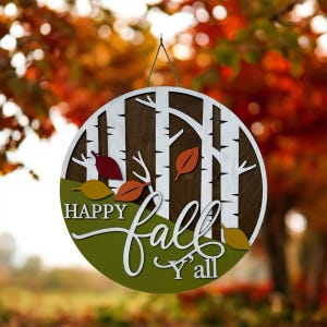 May include: A circular wooden wall sign featuring a white birch tree forest design against a dark brown background. The sign includes a green foreground with scattered red, orange, and yellow leaves. The text reads Happy fall Y'all in white.