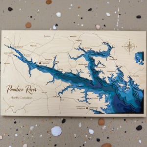 May include: A wooden map of the Pamlico River in North Carolina, featuring layered blue depths. The map includes place names like Washington, Belhaven, and Aurora, with a compass rose in the upper right corner. The text "Pamlico River North Carolina" is visible.