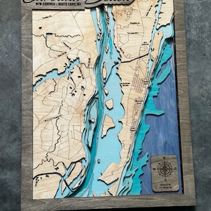 Puede incluir: Un mapa de madera de Carolina Beach, Carolina del Norte, con detalles en capas azules y beige que representan la costa y las vías fluviales. El mapa está enmarcado en madera gris e incluye el texto "Carolina Beach" en la parte superior. Una rosa de los vientos está en la esquina inferior derecha.