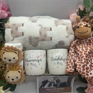May include: A baby gift box with a white blanket patterned with brown shapes, two mugs reading "Mummy's Mug" and "Daddy's Mug", a giraffe soft toy, two lion-shaped baby socks, and a photo card. The box is surrounded by pink flowers.