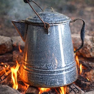 May include: A gray speckled enamel coffee percolator with a metal handle, a spout, and a lid. The pot features a wire bail handle with a small wooden knob at the top. It is positioned directly over an open fire with visible flames and burning wood.