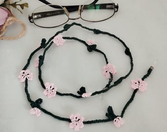 May include: A dark green crocheted eyeglass chain featuring small pink flower embellishments. The chain is designed to hold spectacles, with a pair of glasses resting on the surface. The floral design adds a touch of elegance.