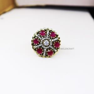 May include: A gold ring with a circular design featuring red and white stones. The ring has a decorative border and a central white stone surrounded by smaller red stones.