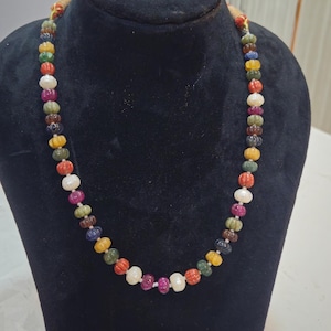 May include: A colorful beaded necklace displayed on a black bust. The necklace features a variety of gemstone beads in shades of red, yellow, green, blue, and white. The beads have a textured, ridged design.