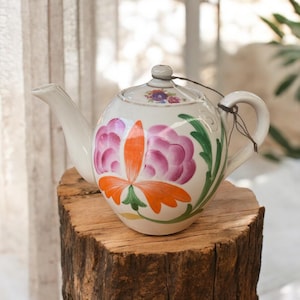 May include: A white teapot with a floral design. The teapot has a pink, orange, and green floral pattern. The teapot is sitting on a wooden stump.