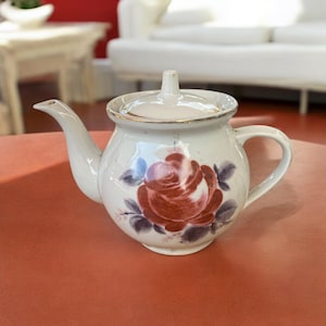 May include: A white porcelain teapot with a floral design. The teapot has a lid and a handle. The floral design features a red rose with purple leaves.
