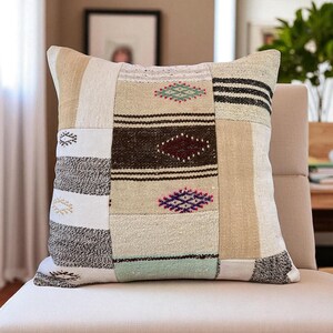 May include: Square patchwork cushion in neutral tones of beige, cream, and brown. It features geometric patterns and stripes in black, green, and purple. The cushion is placed on a beige chair. Measurements are not available.