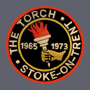 May include: A circular, red-bordered patch with a black background. The text "THE TORCH" and "STOKE-ON-TRENT" are in a white, cursive font. The text "1965" and "1973" are in a white, sans-serif font. A yellow hand holds a red and white torch.
