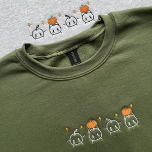 May include: A light gray crewneck sweatshirt and an olive green crewneck sweatshirt, both featuring embroidered designs of four small white cartoon characters, with two of them holding orange pumpkins and surrounded by small orange star accents.