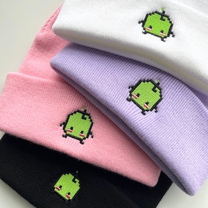 May include: Four knit beanies in different colors, white, purple, pink, and black, with a green pixelated character embroidered on each.