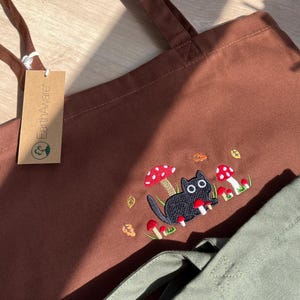 以下が含まれることがあります： 黒猫とキノコの刺繍デザインが施された茶色のトートバッグ。猫は大きな白い目を持ち、赤と白のキノコと秋の葉に囲まれています。バッグには長い茶色のハンドルと「EarthAware」と書かれたタグが付いています。