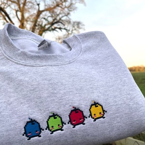 May include: A gray sweatshirt with a row of five embroidered pixelated characters. The characters are blue, green, red, yellow, and yellow. Each character has a small red flame on top of its head.