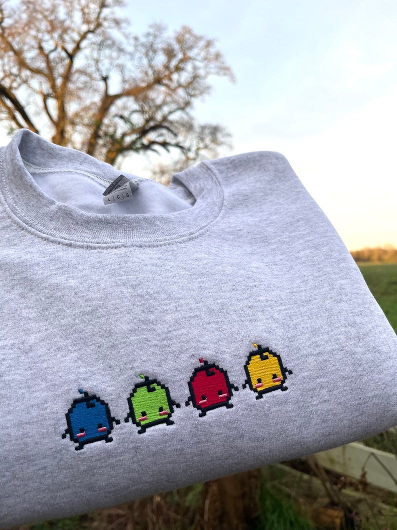 May include: A gray sweatshirt with a row of five embroidered pixelated characters. The characters are blue, green, red, yellow, and yellow. Each character has a small red flame on top of its head.