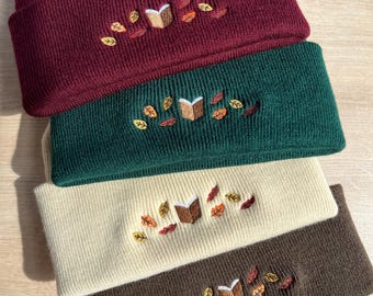 Pode incluir: Quatro gorros de malha em bordô, verde floresta, creme e castanho. Cada gorro apresenta um design bordado de um livro aberto com folhas de outono. Os gorros estão dobrados e empilhados, mostrando as diferentes cores e designs.