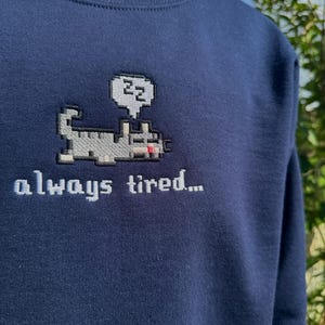 May include: A navy blue sweatshirt with a pixelated cat design and the text "always tired..." embroidered on the front. The cat is wearing a speech bubble with the letters "zz" inside.