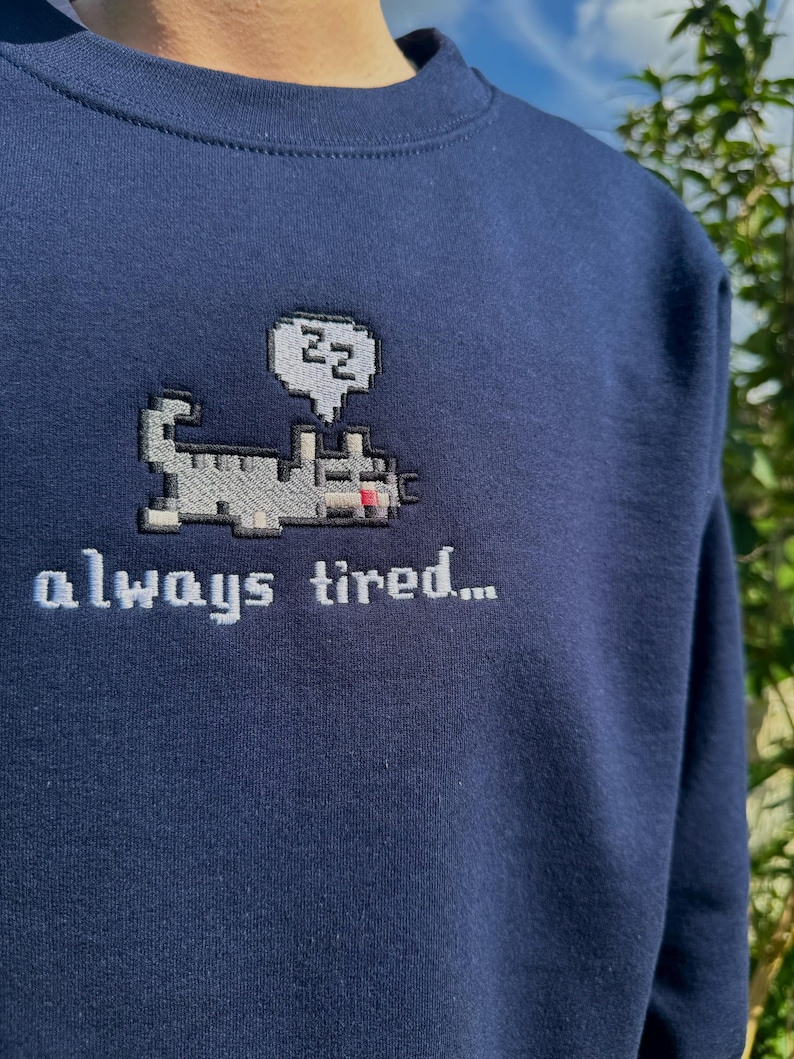 May include: A navy blue sweatshirt with a pixelated cat design and the text "always tired..." embroidered on the front. The cat is wearing a speech bubble with the letters "zz" inside.