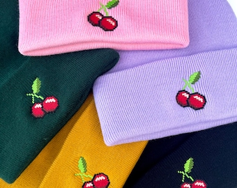 May include: A selection of beanies in various colours, including pink, green, yellow, purple, and navy. Each beanie has a pixelated cherry embroidery on the folded brim. The beanies are made of a soft, ribbed knit material.