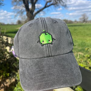 May include: A charcoal gray baseball cap featuring a pixelated green alien design with a pink mouth and black eyes. The cap has a curved brim and an adjustable strap. The background shows a green field and a blue sky.
