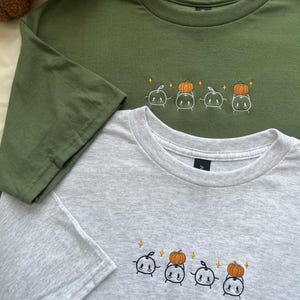 May include: An olive green t-shirt and a heather grey t-shirt, each featuring an embroidered design of four small white ghost-like figures. Two of the figures are holding orange pumpkins above their heads, accented by small yellow star shapes.
