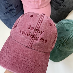 Puede incluir: Una colección de cinco gorras de béisbol en varios colores, incluyendo granate, rosa, gris oscuro, azul y verde. Cada gorra presenta un acabado de tela desgastada y el texto bordado "in my reading era" en el panel frontal.