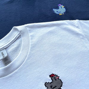 May include: Two t-shirts, one navy blue and one white, each with a pixelated embroidered chicken design. The chicken on the white shirt is wearing a red hat.