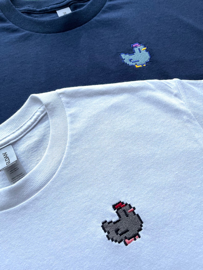 May include: Two t-shirts, one navy blue and one white, each with a pixelated embroidered chicken design. The chicken on the white shirt is wearing a red hat.