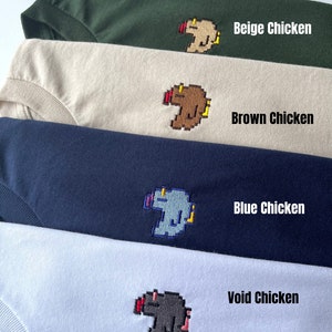 May include: Four t-shirts with different colored pixelated chicken designs. The shirts are stacked on top of each other. The designs are beige, brown, blue, and gray. The text on the shirts reads "Beige Chicken", "Brown Chicken", "Blue Chicken", and "Void Chicken".