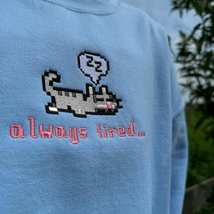 May include: A light blue sweatshirt with a pixelated cat design and the text "always tired..." embroidered in pink.