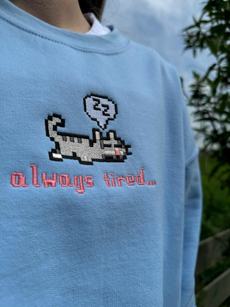 May include: A light blue sweatshirt with a pixelated cat design and the text "always tired..." embroidered in pink.