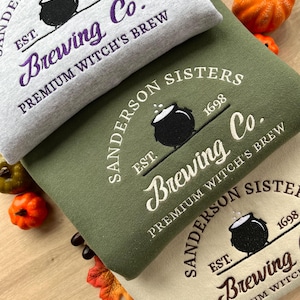 Può includere: Tre felpe con cappuccio impilate l'una sull'altra. La felpa superiore è grigia, quella di mezzo è verde e quella inferiore è beige. Ogni felpa ha un calderone nero e il testo "Sanderson Sisters Est. 1698 Brewing Co. Premium Witch's Brew" ricamato sul davanti.