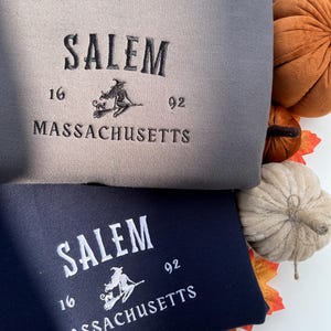 May include: Two sweatshirts, one grey and one navy blue, are displayed. Both feature the word "SALEM" arched across the top, with "16" and "92" on either side of a witch on a broom. The word "MASSACHUSETTS" is below. Decorative pumpkins and autumn leaves surround the sweatshirts.