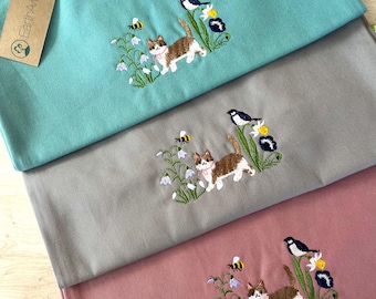 Puede incluir: Tres bolsas de tela de diferentes colores. Las bolsas están apiladas una encima de la otra. La bolsa de arriba es turquesa, la del medio es gris y la de abajo es rosa. Cada bolsa tiene un diseño bordado de un gato caminando por un campo de flores con una abeja y un pájaro. Las bolsas tienen una etiqueta que dice "EarthAware".