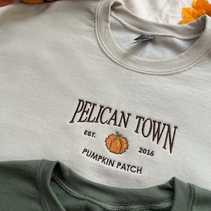 May include: A beige crewneck sweatshirt featuring dark brown embroidered text that reads PELICAN TOWN EST. 2016 PUMPKIN PATCH with a pixelated orange pumpkin graphic in the centre. A dark green sweatshirt is partially visible at the bottom edge.