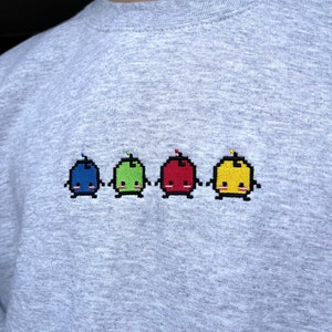 May include: A gray sweatshirt with a row of five embroidered pixelated characters. The characters are blue, green, red, yellow, and yellow. Each character has a small red, green, or yellow antenna on top of its head.