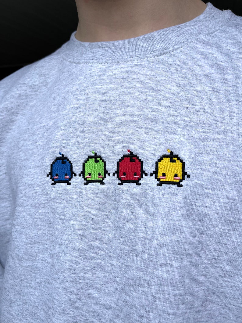 May include: A gray sweatshirt with a row of five embroidered pixelated characters. The characters are blue, green, red, yellow, and yellow. Each character has a small red, green, or yellow antenna on top of its head.
