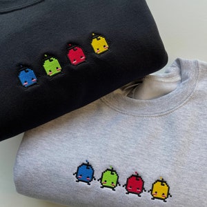 May include: Two sweatshirts with embroidered pixelated characters. The top sweatshirt is black with five characters in blue, green, red, yellow, and yellow. The bottom sweatshirt is gray with five characters in blue, green, red, and yellow.