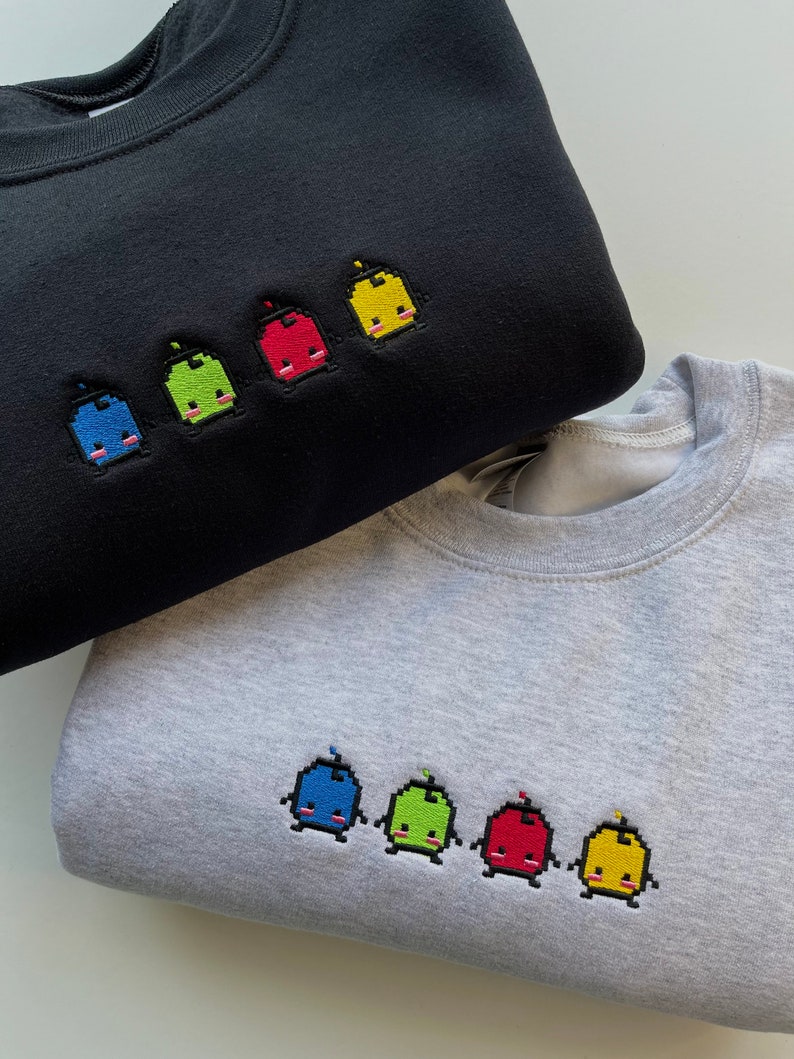 May include: Two sweatshirts with embroidered pixelated characters. The top sweatshirt is black with five characters in blue, green, red, yellow, and yellow. The bottom sweatshirt is gray with five characters in blue, green, red, and yellow.