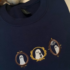 May include: Navy blue sweatshirt with three embroidered ghost portraits in decorative frames. The ghosts are white, with brown and gold frame details. A small pumpkin is visible in the background.