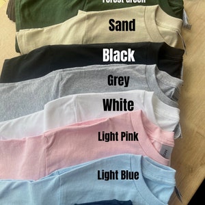 May include: A stack of t-shirts in various colors, including chocolate, forest green, sand, black, grey, white, light pink, light blue, and navy.