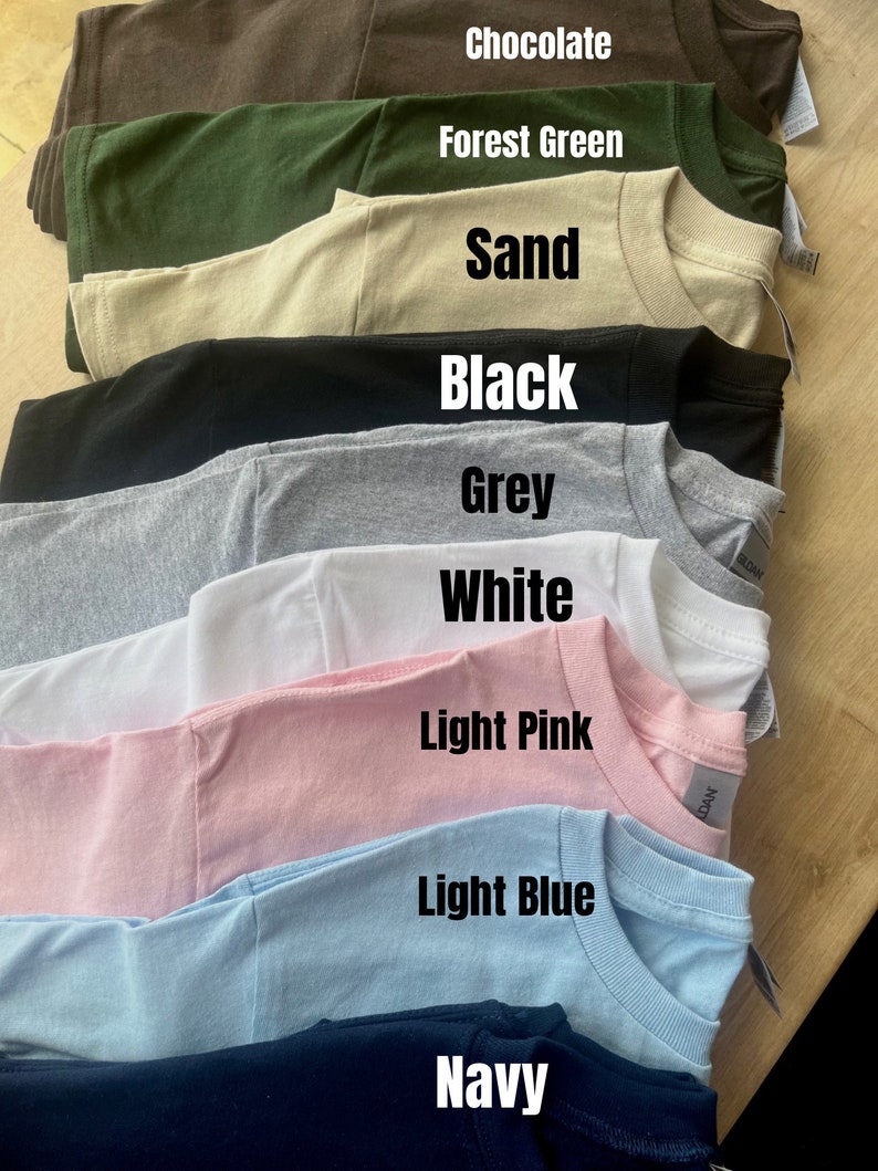 May include: A stack of t-shirts in various colors, including chocolate, forest green, sand, black, grey, white, light pink, light blue, and navy.