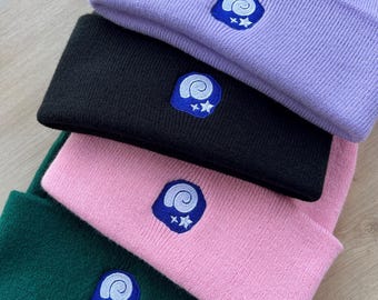 May include: A stack of four knit beanies in shades of purple, black, pink, and green. Each beanie features a blue embroidered patch with a white spiral design and two white stars. The beanies are folded and stacked on top of each other.