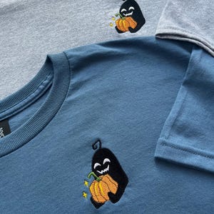 May include: A light grey t-shirt and a blue t-shirt, each featuring an embroidered design of a black ghost-like figure holding an orange pumpkin with a green stem. The ghost has a wide, white, jagged-toothed smile and small yellow star accents.